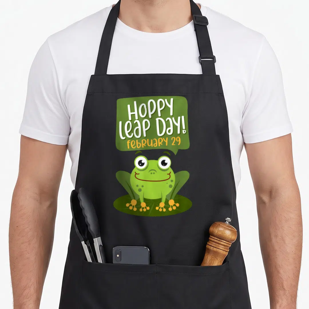 Hoppy frog aprons - Cartoon green frog on lily pad with Hoppy Leap Day text in speech bubble — shown on a black cotton canvas apron against a white background