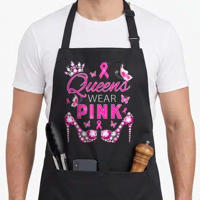 Queen ribbon apron - Stacked text Queens WEAR PINK with jeweled crown, butterflies, and diamond-encrusted high heels — shown on a black cotton canvas apron against a white background