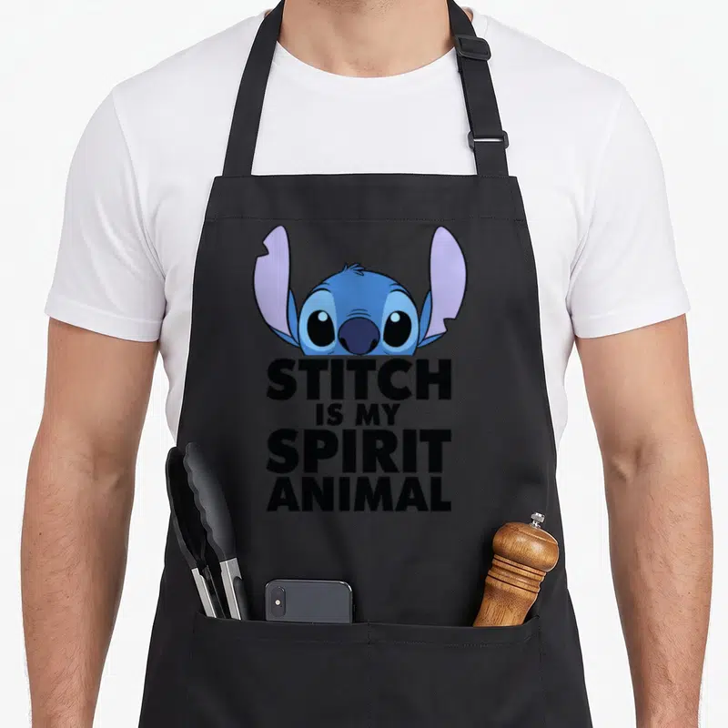 Critter apron - Blue cartoon head above four lines of black text reading Stitch Is My Spirit Animal — shown on a black cotton canvas apron against a white background