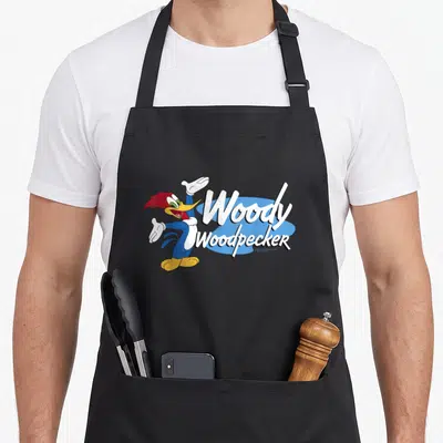 Bold woodpecker apron - Woody Woodpecker cartoon character in red blue white yellow with stacked white text on light — shown on a black cotton canvas apron against a white background