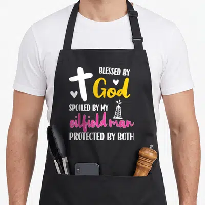 Oilfield husband wife aprons - Cross and oil derrick graphic with text Blessed by God Spoiled by My Oilfield Man — shown on a black cotton canvas apron against a white background