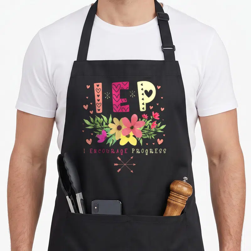 Special education teacher apron - IEP letters above pink yellow and white flowers with crossed arrows and text — shown on a black cotton canvas apron against a white background