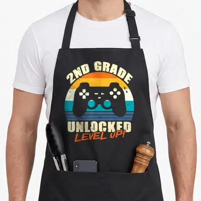Unlocked 2nd apron - Grey game controller over orange turquoise beige striped sunset circle with 2nd grade text — shown on a black cotton canvas apron against a white background