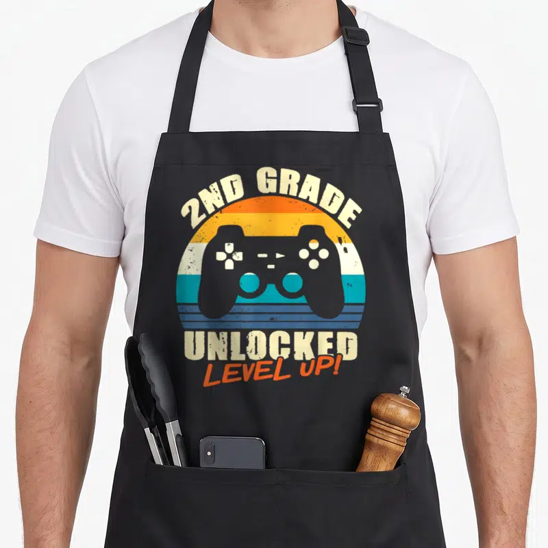 Unlocked 2nd apron - Grey game controller over orange turquoise beige striped sunset circle with 2nd grade text — shown on a black cotton canvas apron against a white background