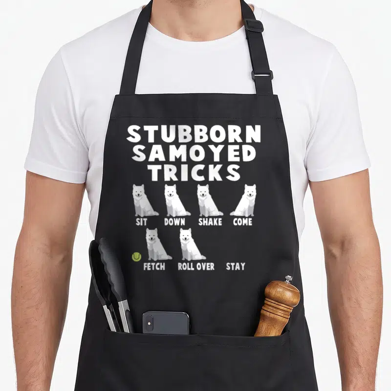 Stubborn samoyed apron - Cartoon Samoyeds listed under stubborn tricks with a lime green tennis ball — shown on a black cotton canvas apron against a white background