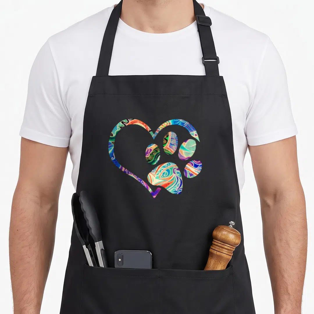 Animal paw print apron - Heart shape enclosing a paw print filled with colorful psychedelic swirls in blue, green — shown on a black cotton canvas apron against a white background