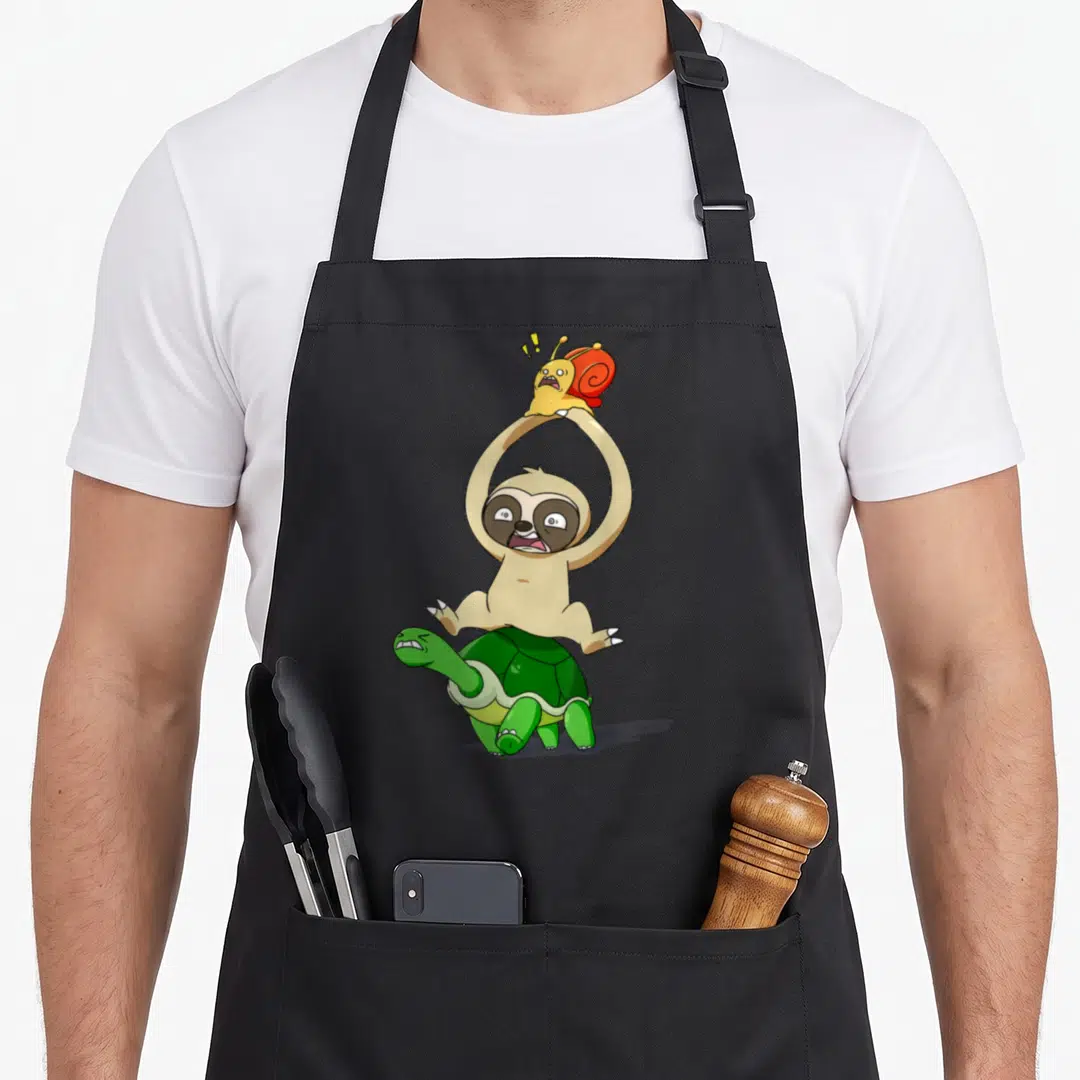 Marathon sloth apron - Cartoon beige sloth on green turtle holding yellow snail with red shell — shown on a black cotton canvas apron against a white background