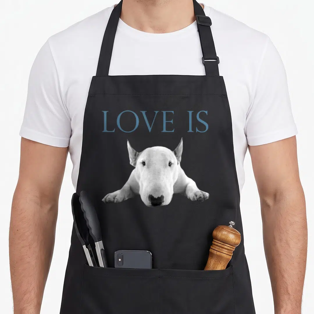 Funny bull terrier loves apron - Blue-grey LOVE IS text above a white bull terrier dog lying down on grey background — shown on a black cotton canvas apron against a white background