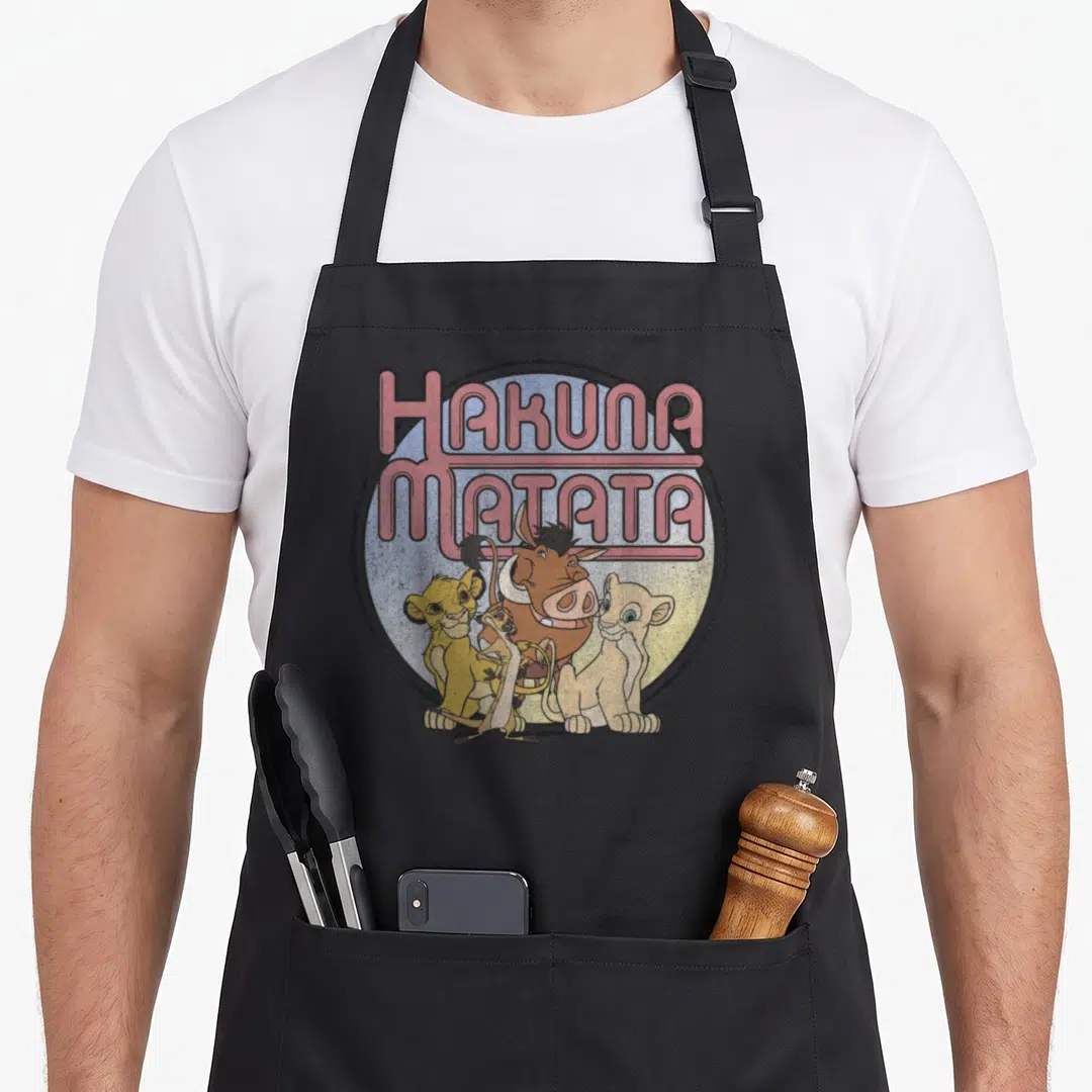 Hakuna lion apron - Retro pink HAKUNA MATATA text above four cartoon animals including Pumbaa in a circle — shown on a black cotton canvas apron against a white background