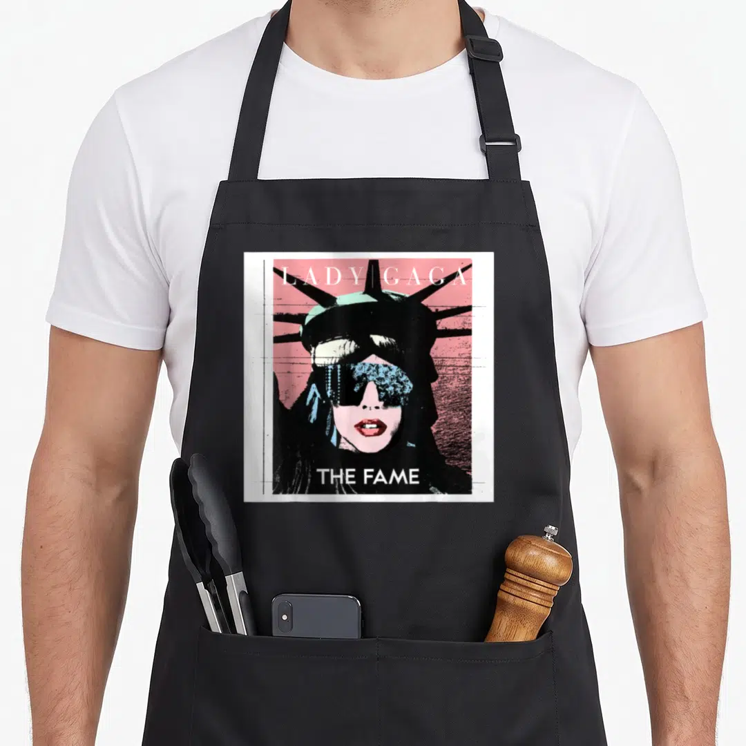 Gaga liberty apron - Lady Gaga The Fame graphic with woman head between text lines in pink black white and green — shown on a black cotton canvas apron against a white background