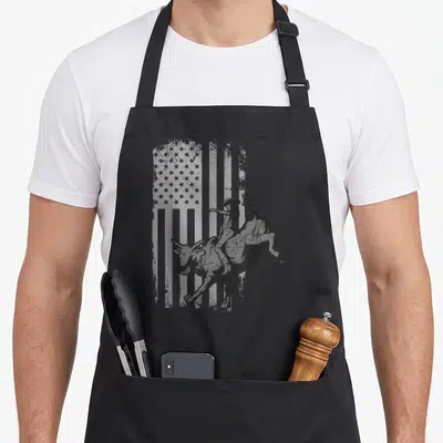 Bucking american flag apron - Distressed black and grey bull rider silhouette overlaid on a faded American flag pattern — shown on a black cotton canvas apron against a white background