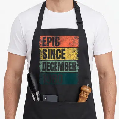 Epic december 40th birthday apron - Grey EPIC SINCE DECEMBER 1980 text on orange yellow green and teal distressed bars — shown on a black cotton canvas apron against a white background