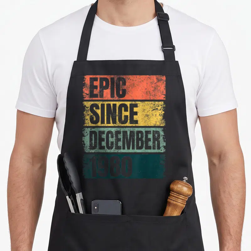 Epic december 40th birthday apron - Grey EPIC SINCE DECEMBER 1980 text on orange yellow green and teal distressed bars — shown on a black cotton canvas apron against a white background