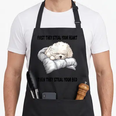 Sleeping bichon frise apron - Cartoon white dog sleeping on grey pillow with black text quote about stealing hearts — shown on a black cotton canvas apron against a white background