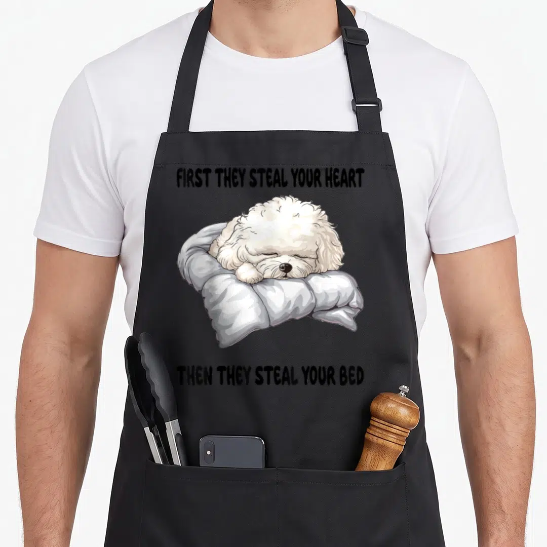 Sleeping bichon frise apron - Cartoon white dog sleeping on grey pillow with black text quote about stealing hearts — shown on a black cotton canvas apron against a white background