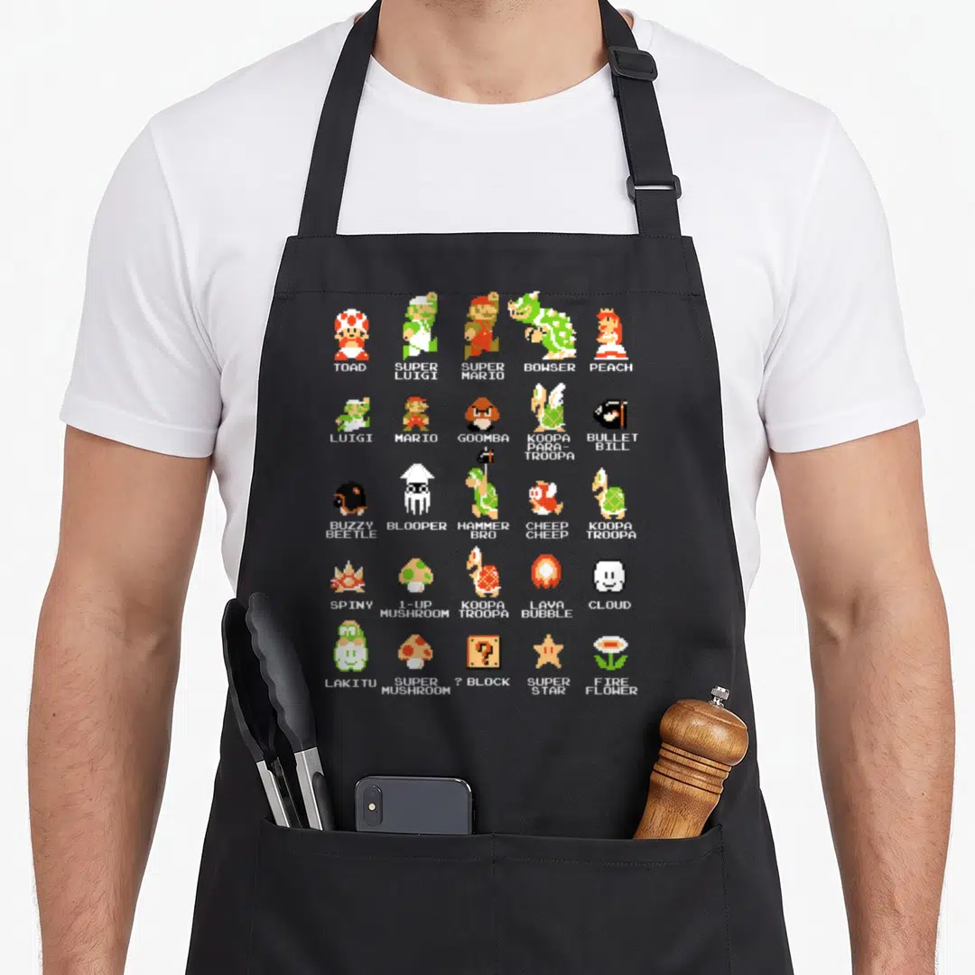 Bit boy pixel apron - Grid of pixel art characters including MARIO, PEACH, BOWSER, and items like SUPER STAR labeled — shown on a black cotton canvas apron against a white background