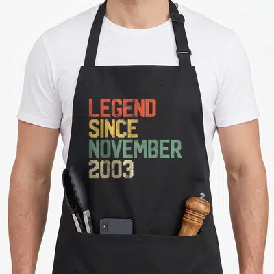 Legend november birthday apron - Stacked distressed text in orange yellow green and beige reading LEGEND SINCE NOVEMBER — shown on a black cotton canvas apron against a white background