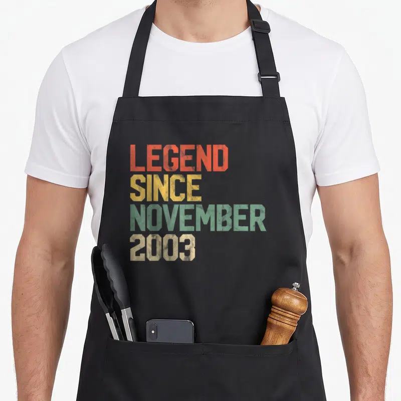 Legend november birthday apron - Stacked distressed text in orange yellow green and beige reading LEGEND SINCE NOVEMBER — shown on a black cotton canvas apron against a white background