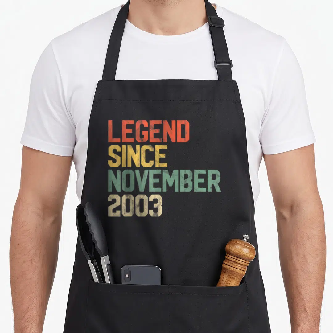 Legend november birthday apron - Stacked distressed text in orange yellow green and beige reading LEGEND SINCE NOVEMBER — shown on a black cotton canvas apron against a white background