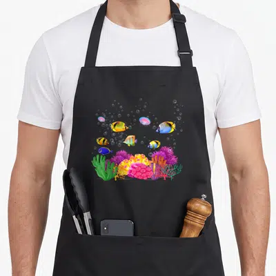 Colourful tropical print aprons - Cartoon coral reef with fish, jellyfish, and bubbles in yellow, blue, pink, purple — shown on a black cotton canvas apron against a white background