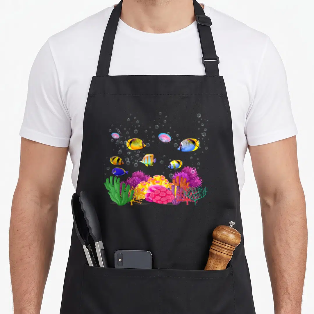 Colourful tropical print aprons - Cartoon coral reef with fish, jellyfish, and bubbles in yellow, blue, pink, purple — shown on a black cotton canvas apron against a white background