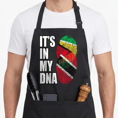 Trinidadian guyanese heritage aprons - IT'S IN MY DNA text beside a fingerprint striped with green yellow red white — shown on a black cotton canvas apron against a white background