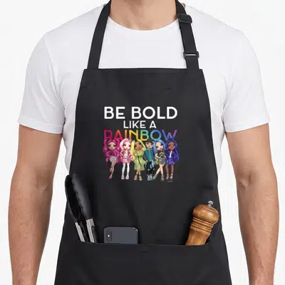 Bold rainbow apron - BE BOLD LIKE A RAINBOW text above six cartoon fashion dolls in white, pink, blue, yellow — shown on a black cotton canvas apron against a white background