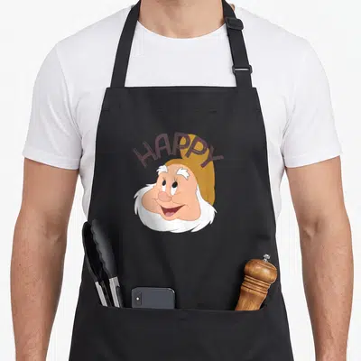 Face snow white apron - Happy dwarf face with arched HAPPY text in peach and brown cartoon style — shown on a black cotton canvas apron against a white background