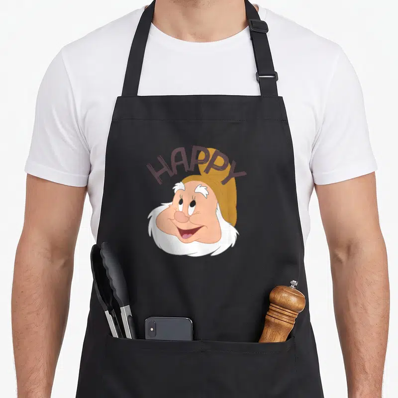 Face snow white apron - Happy dwarf face with arched HAPPY text in peach and brown cartoon style — shown on a black cotton canvas apron against a white background