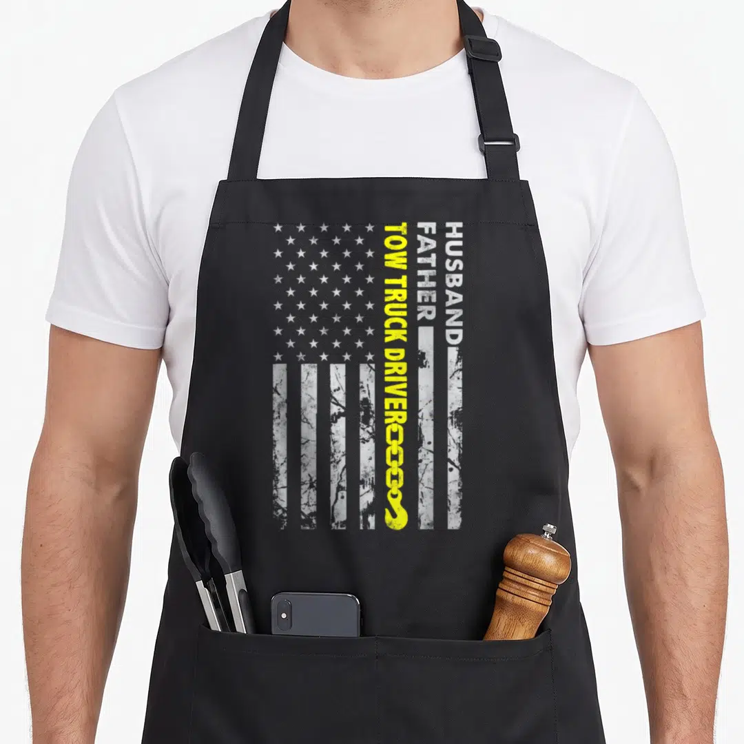 Husband american flag apron - Vertical HUSBAND FATHER TOW TRUCK DRIVER text with tow hook on distressed white — shown on a black cotton canvas apron against a white background
