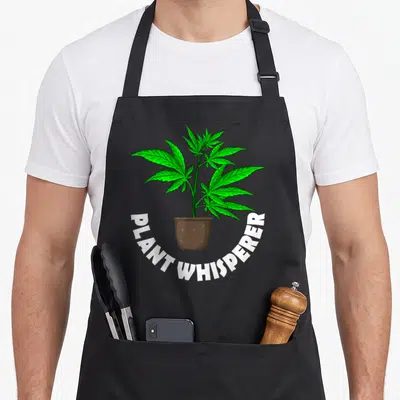 Weed hemp apron - Cartoon green cannabis plant in brown pot with PLANT WHISPERER text arching below — shown on a black cotton canvas apron against a white background
