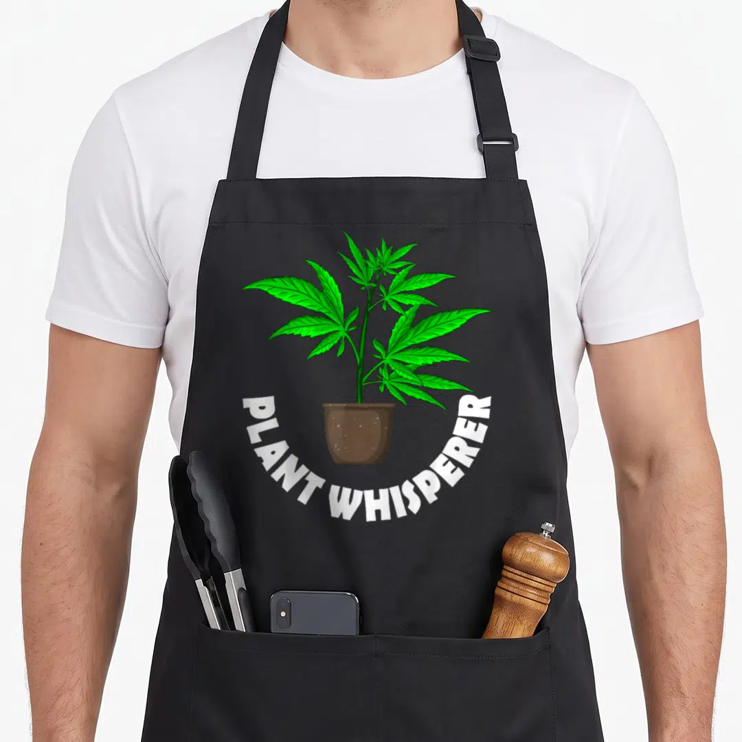 Weed hemp apron - Cartoon green cannabis plant in brown pot with PLANT WHISPERER text arching below — shown on a black cotton canvas apron against a white background