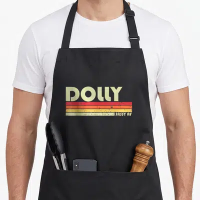 Dolly birthday apron - Retro DOLLY text above red orange yellow beige stripes with SASSY AF label — shown on a black cotton canvas apron against a white background
