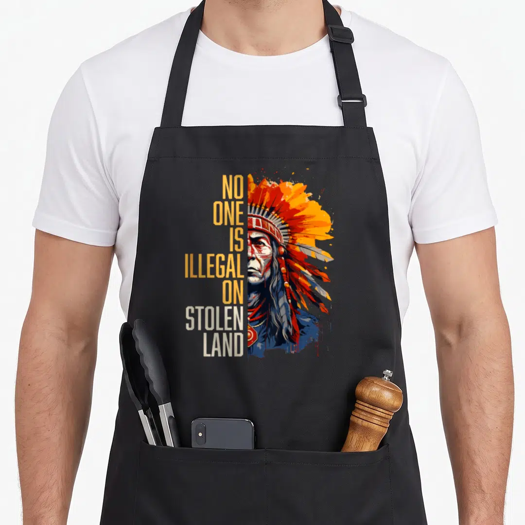 Illegal indigenous apron - Stacked text No One Is Illegal On Stolen Land beside a realistic Native American face — shown on a black cotton canvas apron against a white background
