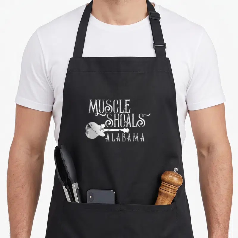 Muscle alabama apron - White distressed text MUSCLE SHOALS ALABAMA with horizontal guitar silhouette overlapping — shown on a black cotton canvas apron against a white background