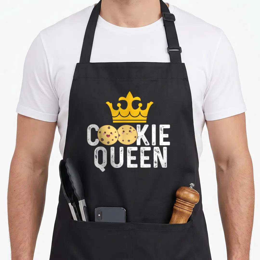 Cookie baker apron - Yellow crown above white COOKIE QUEEN text with two smiling tan cookies replacing O letters — shown on a black cotton canvas apron against a white background