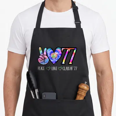 Class 50th tie-dye apron - Tie-dye peace sign, heart, 77 graphic with PEACE LOVE CLASS OF '77 text and two small hearts — shown on a black cotton canvas apron against a white background