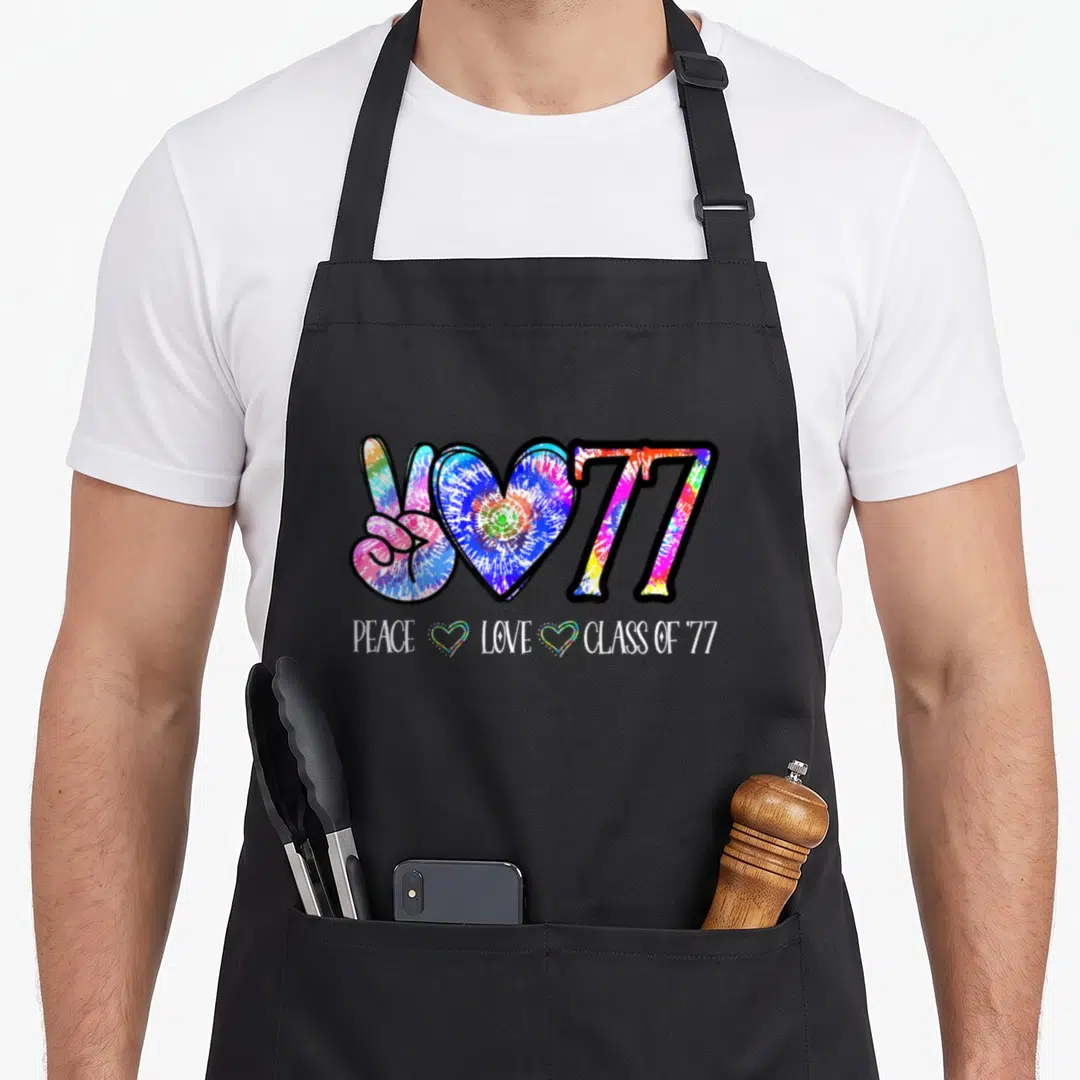 Class 50th tie-dye apron - Tie-dye peace sign, heart, 77 graphic with PEACE LOVE CLASS OF '77 text and two small hearts — shown on a black cotton canvas apron against a white background