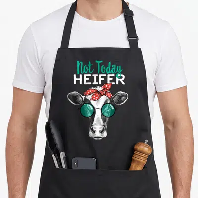 Today country apron - Cartoon cow wearing green sunglasses and red polka dot bandana with Not Today Heifer text — shown on a black cotton canvas apron against a white background