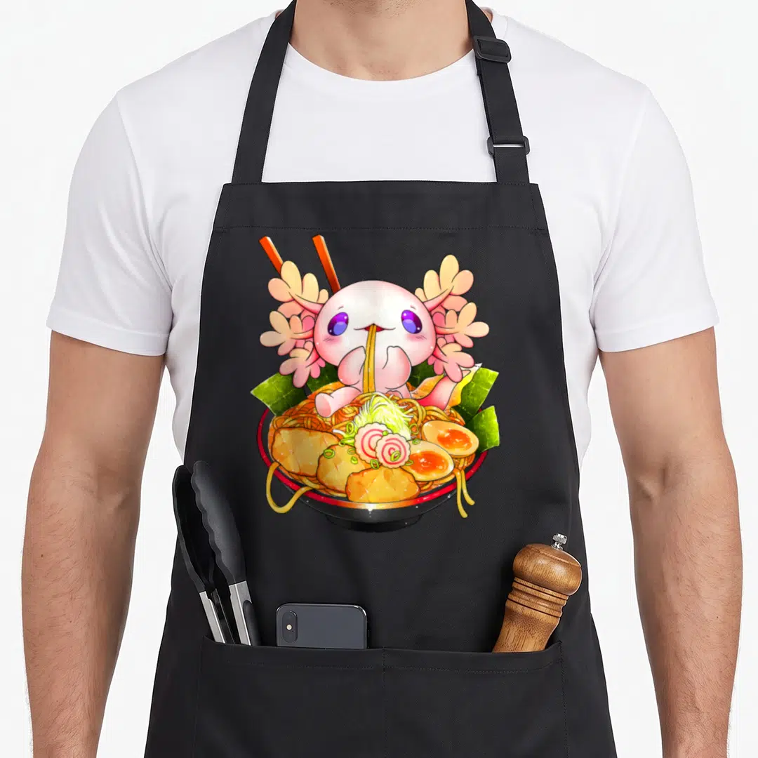 Axolotl ramen anime apron - Pink cartoon axolotl eating ramen noodles in a bowl with chopsticks and green seaweed — shown on a black cotton canvas apron against a white background