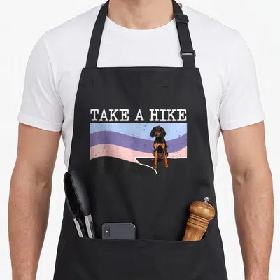 Hike black apron - TAKE A HIKE text above cartoon dachshund on wavy blue purple pink black flag — shown on a black cotton canvas apron against a white background
