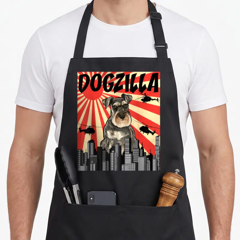 Japanese miniature schnauzer apron - Giant grey dog DOGZILLA behind city skyline with helicopters on red sunburst — shown on a black cotton canvas apron against a white background