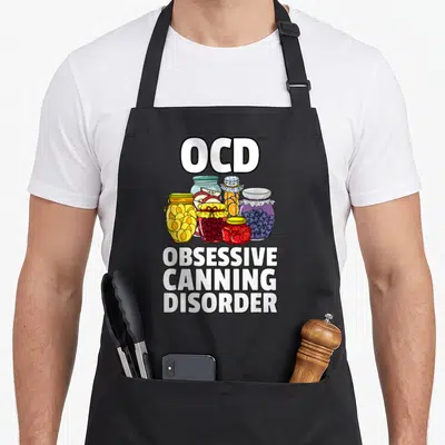 Jar canning apron - Cartoon five fruit jars with OCD OBSESSIVE CANNING DISORDER text in white red blue yellow orange — shown on a black cotton canvas apron against a white background
