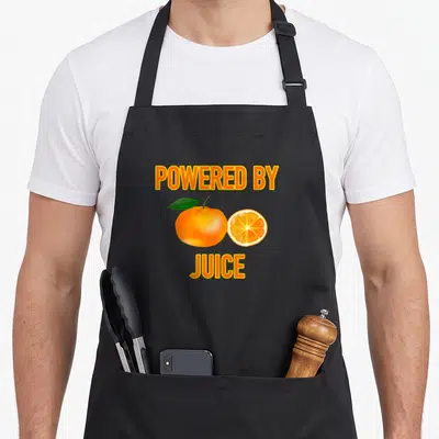Powered funny morning apron - POWERED BY JUICE text above orange and slice with green leaf — shown on a black cotton canvas apron against a white background
