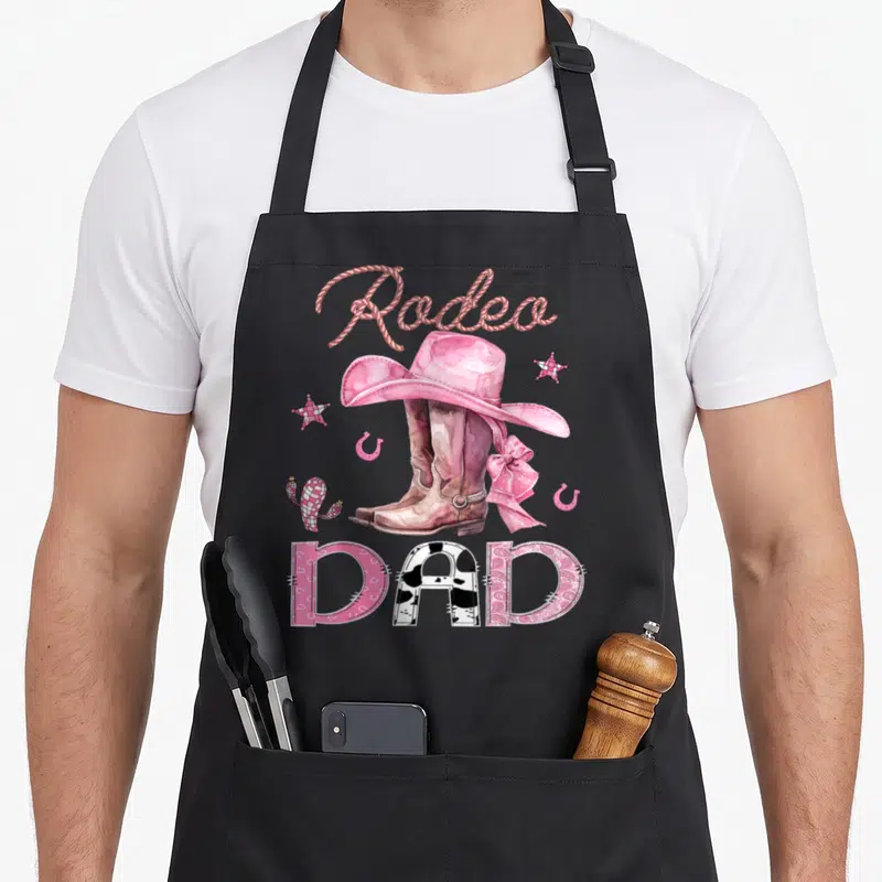 Rodeo boot cowgirl aprons - Rodeo DAD text with pink cowboy boots, hat, cactus, stars, and horseshoes in watercolor — shown on a black cotton canvas apron against a white background