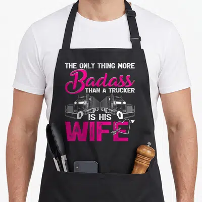 Badass wife apron - Pink and white text with two facing trucks and heart pierced by arrow — shown on a black cotton canvas apron against a white background