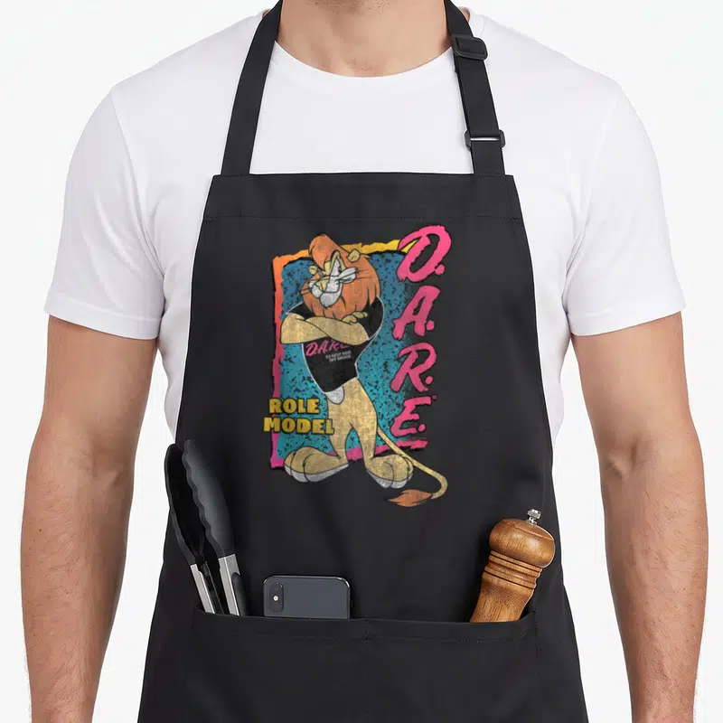 Program lion apron - Cartoon lion with crossed arms and D.A.R.E. text on jagged blue pink background — shown on a black cotton canvas apron against a white background