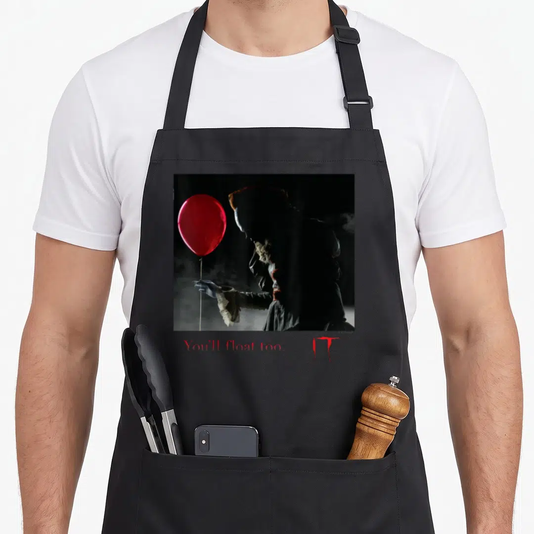 Film balloon apron - Pennywise holding red balloon with You'll float too text below — shown on a black cotton canvas apron against a white background