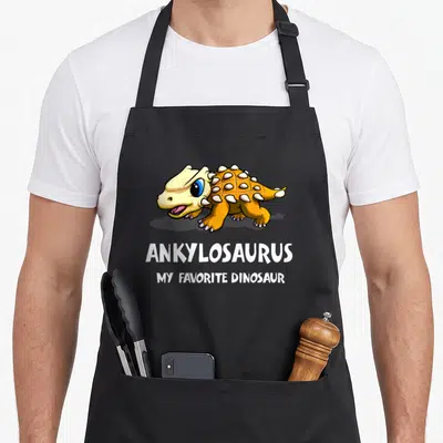 Ankylosaurus dinosaur apron - Cartoon orange ankylosaurus above white text reading Ankylosaurus My Favorite Dinosaur — shown on a black cotton canvas apron against a white background