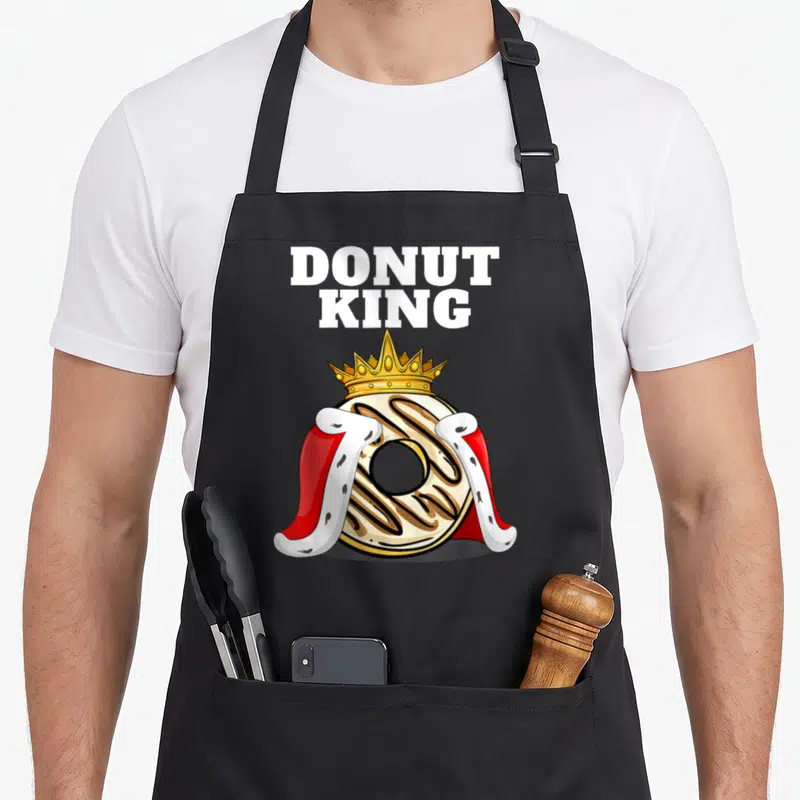 Donut king apron - Cartoon donut with gold crown and red cape under white DONUT KING text — shown on a black cotton canvas apron against a white background